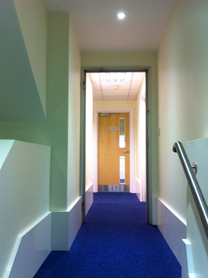 Bright corridor with recessed ceiling lights and blue carpet leading to an office door